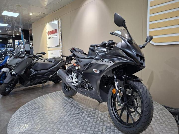 A sleek black Yamaha motorcycle is showcased on a circular display in a dealership, with another motorcycle visible in the background. The setting features neutral walls and a Yamaha sign.