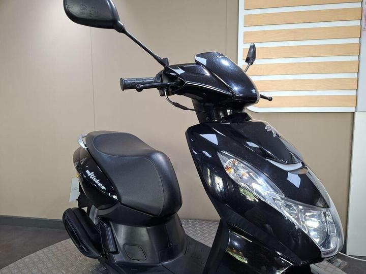 A black scooter displayed in a showroom with a sleek design, featuring a comfortable seat, handlebars, and rearview mirrors. The background is neutral-colored, emphasizing the scooter's features.