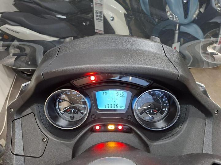 Close-up view of a scooter dashboard displaying speedometer and odometer readings, with illuminated indicator lights, and a digital screen showing time and mileage. The background features blurred scooters.