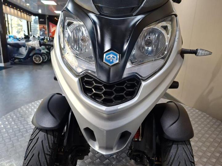 Front view of a silver scooter displaying its headlights and a distinctive logo, set against a blurred background of a vehicle showroom.