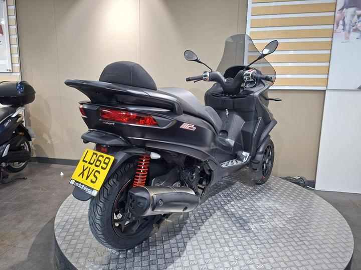 A black three-wheeled scooter displayed on a circular platform, showcasing its rear view with a reflective finish, red suspension springs, and a license plate reading "LD69 XVS." The background features neutral-colored walls and another scooter partially visible on the left.