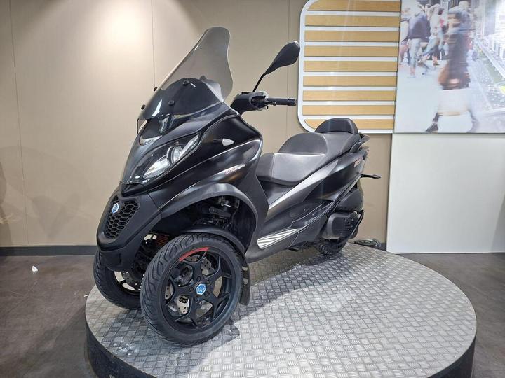 A black three-wheeled scooter displayed on a circular platform, featuring a windshield and modern design elements. The background includes a visible light-colored wall and a screen showing people walking.