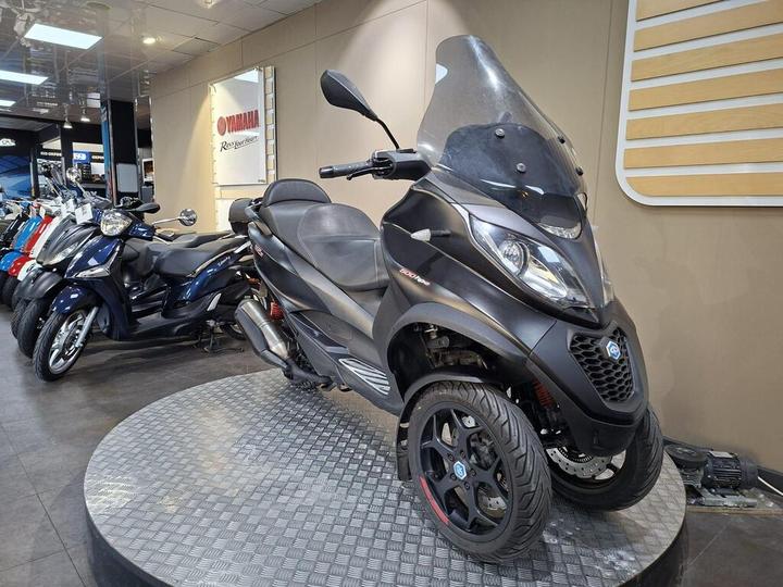 A black three-wheeled scooter displayed on a circular platform in a vehicle showroom, with additional scooters visible in the background and a Yamaha sign on the wall.