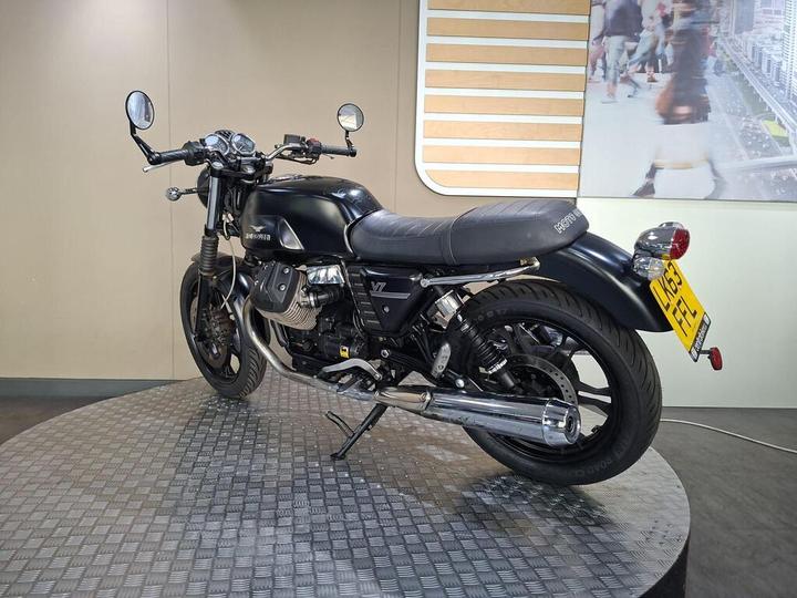 A sleek, black motorcycle with a classic design is displayed on a circular platform. The bike features chrome details, dual mirrors, and a low-slung seat, set against a neutral-colored backdrop.