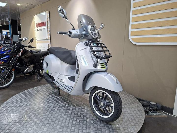 A sleek gray scooter with a chrome headlight and a front grille, displayed on a circular platform in a motorcycle showroom. In the background, a black motorcycle is partially visible alongside a Yamaha sign.