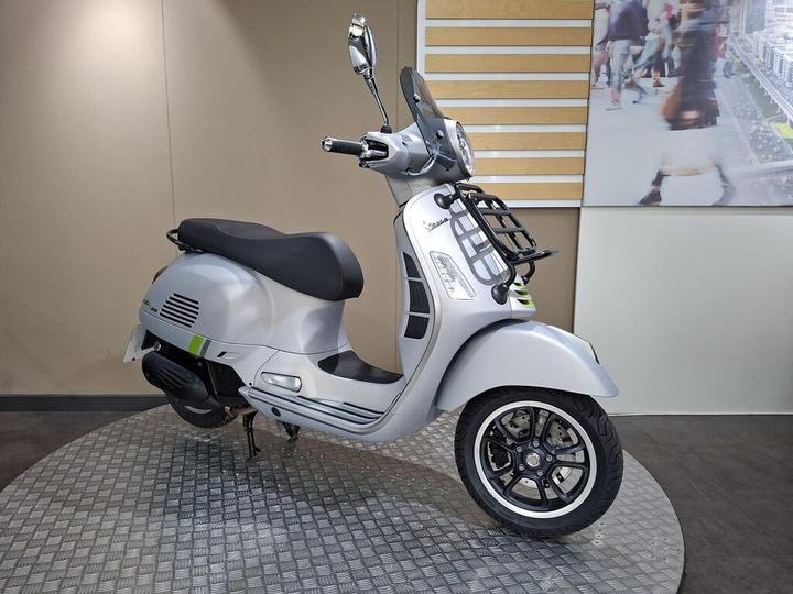 A silver scooter displayed on a textured circular platform, featuring a sleek design, a black seat, and detailed chrome accents, with a softly lit background showcasing a blurred urban setting.