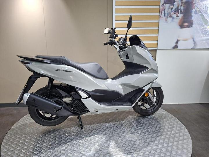 A modern silver scooter displayed on a circular, textured platform against a neutral wall backdrop.