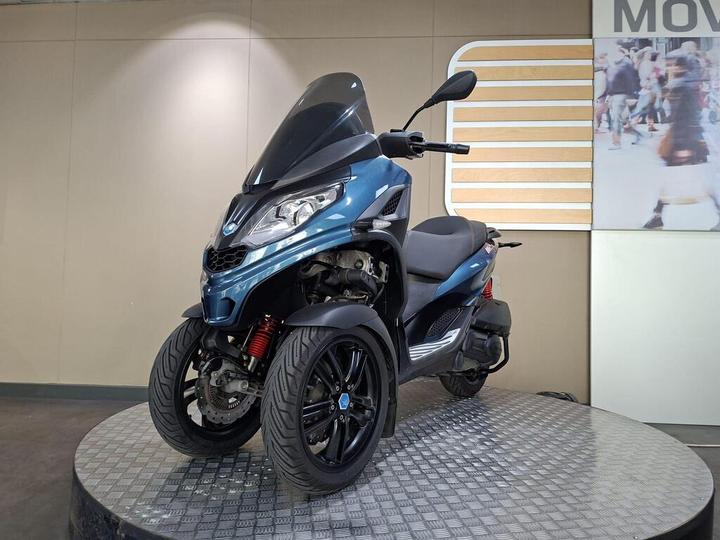 A blue three-wheeled scooter on a circular display stand, featuring a modern design with a large windshield and side mirrors. The background is a neutral-colored wall with a partially visible image of people in motion.