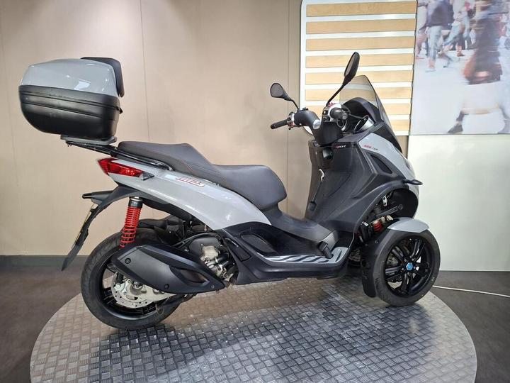 A gray three-wheeled scooter with a top case, showcased in a modern display setting, featuring sporty design elements and red spring suspension.