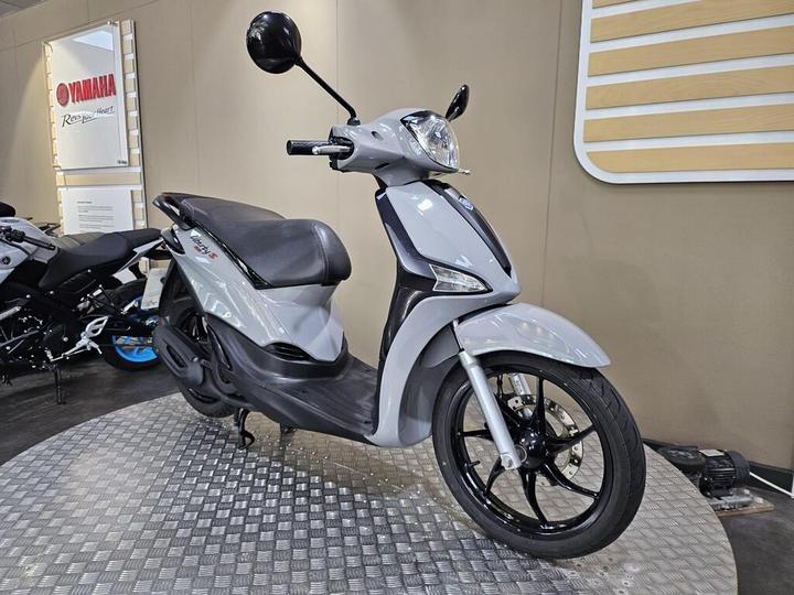 A silver and black scooter displayed on a textured platform in a showroom, with a Yamaha sign visible in the background.