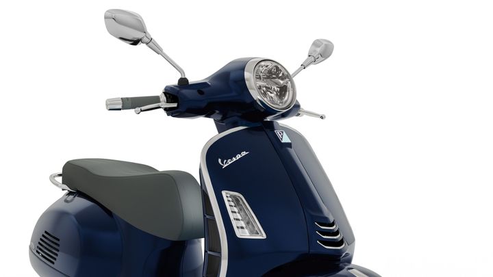 A close-up of a blue Vespa scooter's front, showcasing the handlebars, headlight, and the distinctive body design.