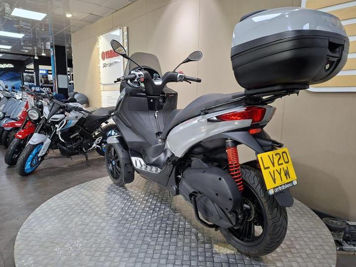 A three-wheeled scooter with a gray body and red suspension springs is displayed in a motorcycle showroom, with various other motorcycles visible in the background. The rear of the scooter features a black top case and a license plate reading "LV20 VYW."