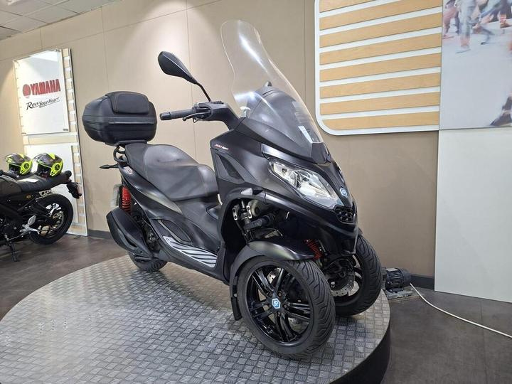 A black scooter with three wheels on a display stand in a showroom, featuring a windshield and a top cargo box, with another scooter in the background.