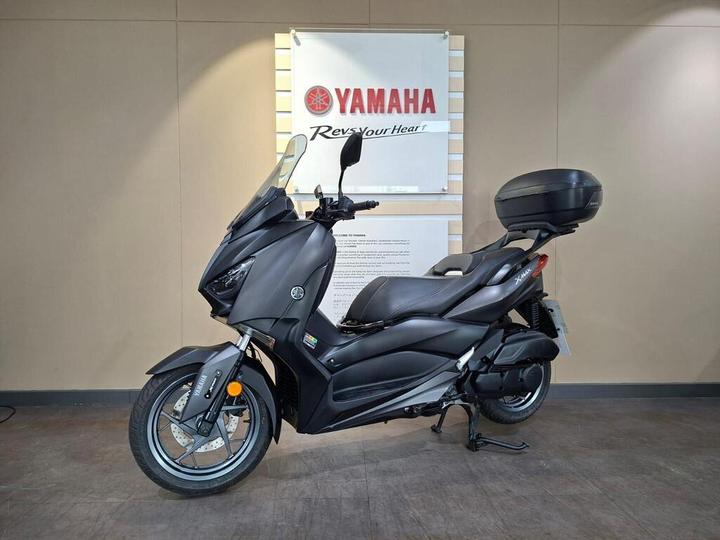 A sleek black Yamaha scooter positioned on a showroom floor, with a promotional sign in the background displaying the Yamaha logo and slogan.