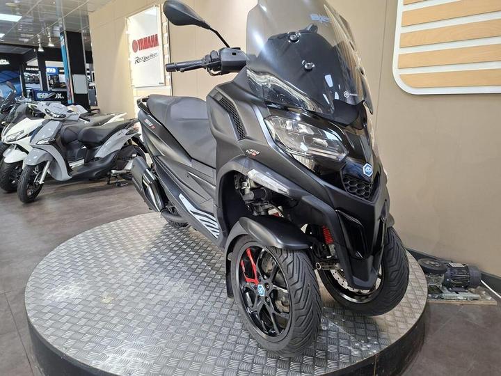 A sleek black three-wheeled scooter displayed on a circular showcase in a dealership, with other scooters visible in the background. The scooter features sporty design elements and red accents on the wheels.
