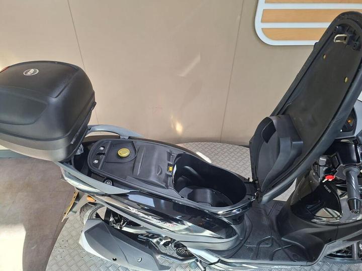 A close-up view of a black scooter with its seat open, revealing the storage compartment and fuel cap beneath, set against a neutral background.