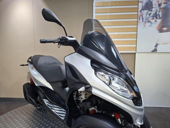 A close-up view of a modern silver scooter with a black front and prominent windshield, displaying intricate design details, parked in an indoor setting with a neutral background.