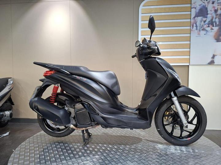 Piaggio Medley S 125 (2023) A side view of a sleek black scooter displayed on a textured surface, with a red shock absorber visible and a light-colored wall in the background.