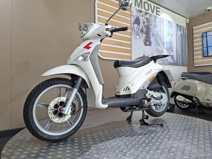 A white scooter with an "L" learner plate displayed on a checkered floor, set against a backdrop of a cityscape in a bright showroom.