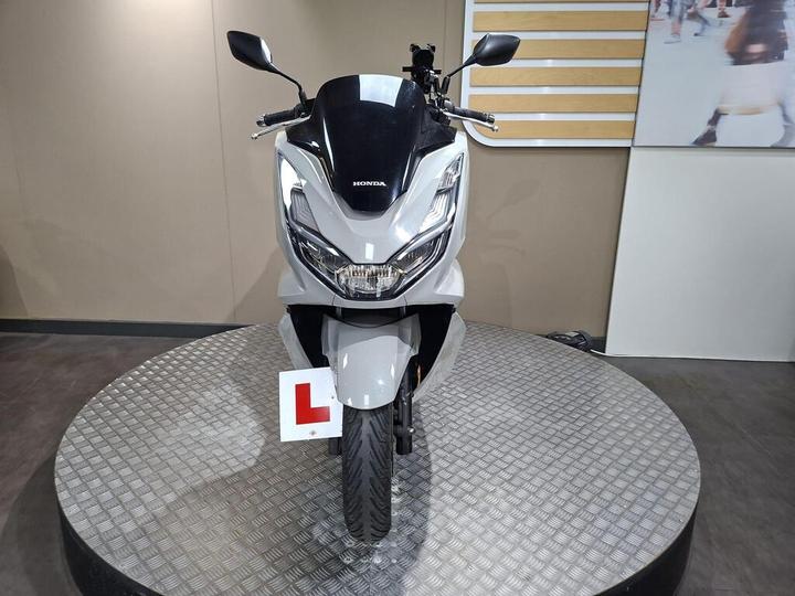 A front view of a silver Honda scooter displayed on a circular platform, featuring a learner plate. The background is neutral with a light-colored wall and a partial view of a screen showing people.