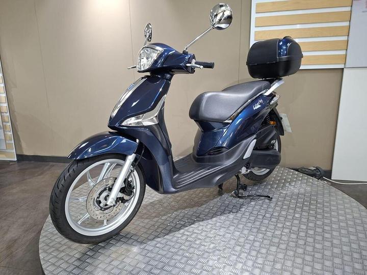 A navy blue scooter with a storage compartment on the back, displayed on a circular platform with a textured surface, set against a neutral-colored wall.