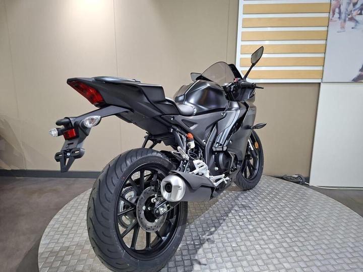 A sleek black motorcycle displayed on a circular metal platform, showcasing its rear and side profile with a prominent exhaust, modern tail light, and sport bike features in a minimalist exhibition space.