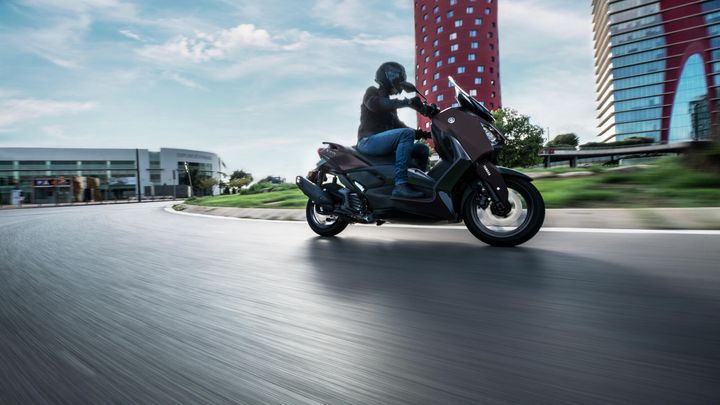 A motorcyclist in a black helmet rides a dark scooter around a curve on a smooth road, with modern buildings and a blue sky in the background.