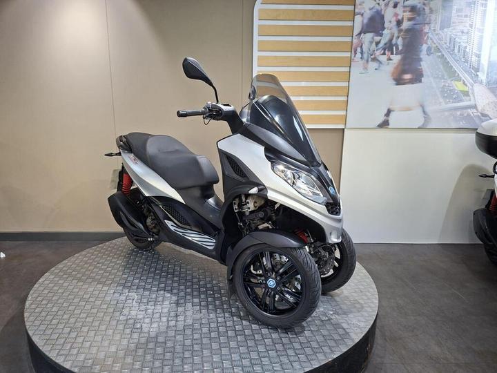 A silver three-wheeled scooter is displayed on a round platform with a textured surface, set against a neutral background featuring a blurred urban scene.