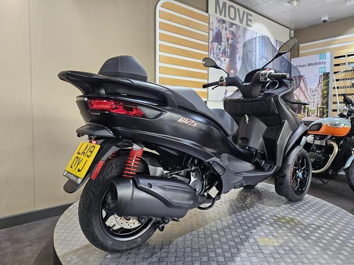 A black scooter, model MP3, displayed in a showroom with a modern interior. The scooter is positioned on a circular metal platform, showcasing its sleek design and distinctive twin front wheels. A backdrop features promotional graphics related to urban mobility.
