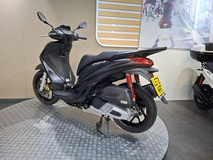 A sleek black scooter displayed on a circular metal platform, showcasing its sporty design and red rear suspension, with a blurred, modern city backdrop.