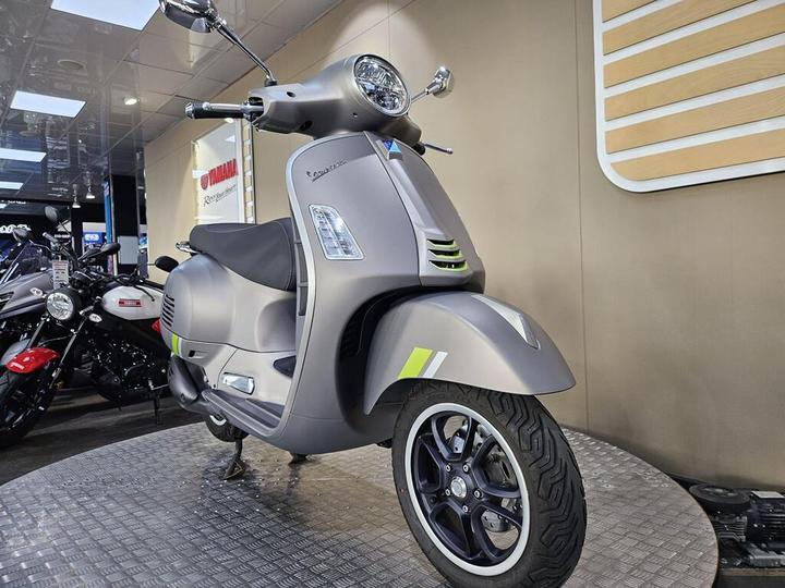 A gray scooter with green accents displayed on a circular platform in a motorcycle showroom, surrounded by other bikes. The scooter features a classic design with a round headlight and sleek body panels.