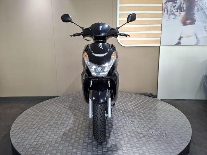 A black scooter positioned on a circular metallic platform, facing forward. The background features a neutral wall and part of an image showing people.