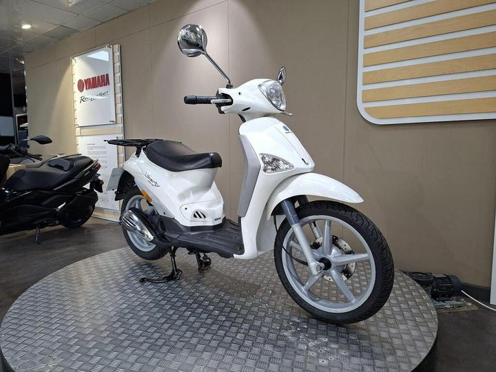 A white scooter displayed on a circular metal platform inside a Yamaha dealership, with a black scooter partially visible in the background.