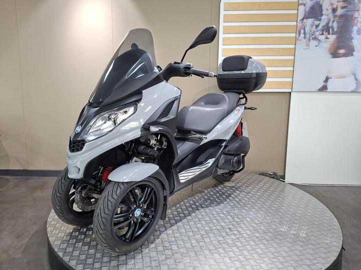 A gray three-wheeled scooter is displayed on a circular platform, featuring a tinted windscreen, modern design, and a storage compartment on the back. The background includes a light-colored wall with a partial image of people walking.