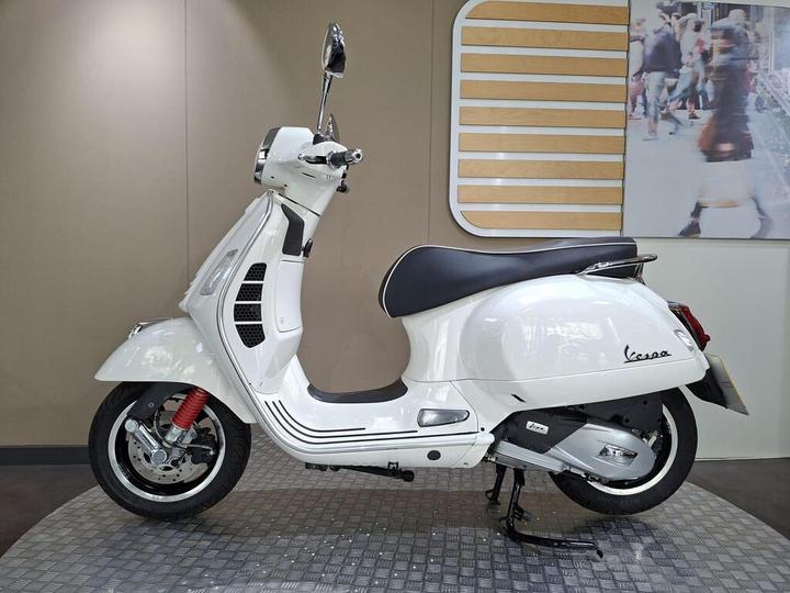 A white Vespa scooter displayed in a showroom, featuring a sleek design with a black seat and red shock absorbers, set against a neutral background.