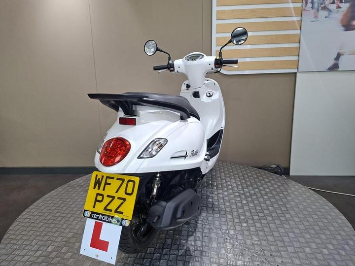 A white scooter displayed on a circular platform, showcasing its rear view, with a visible license plate and a 'learner' driver sign. The background features a neutral-colored wall.