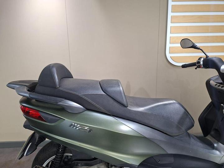 A close-up view of the seat of a green scooter, showcasing its sleek design and textured black covering. The background features a simple beige wall and a wood-paneled accent.