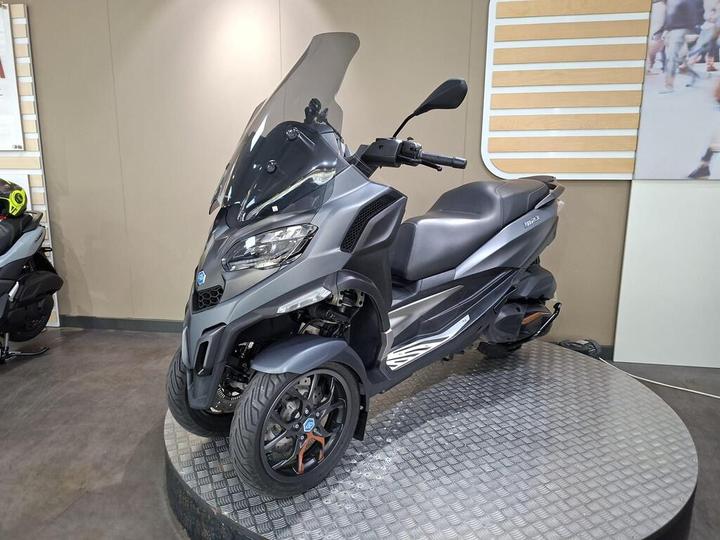 A gray three-wheeled scooter with a windshield displayed on a circular platform in a showroom setting.