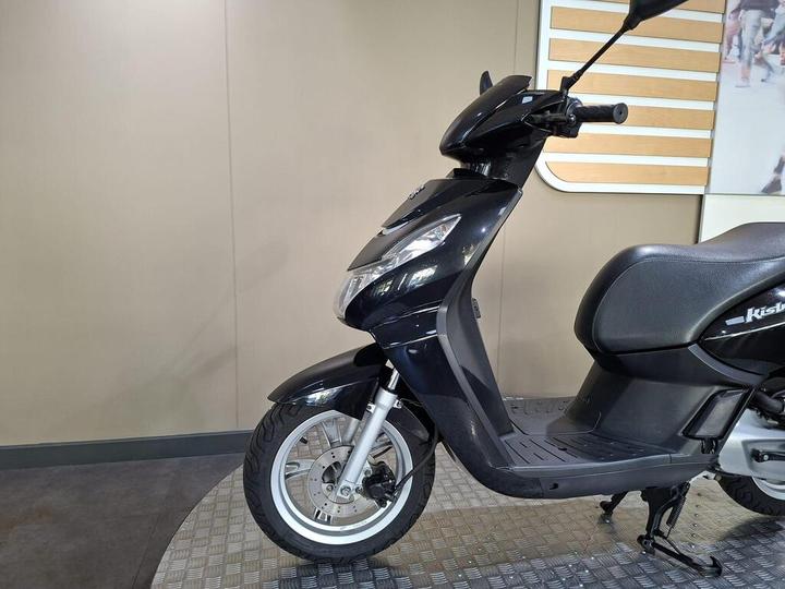 A side view of a black scooter displayed on a textured surface, against a plain wall with a wooden panel in the background.