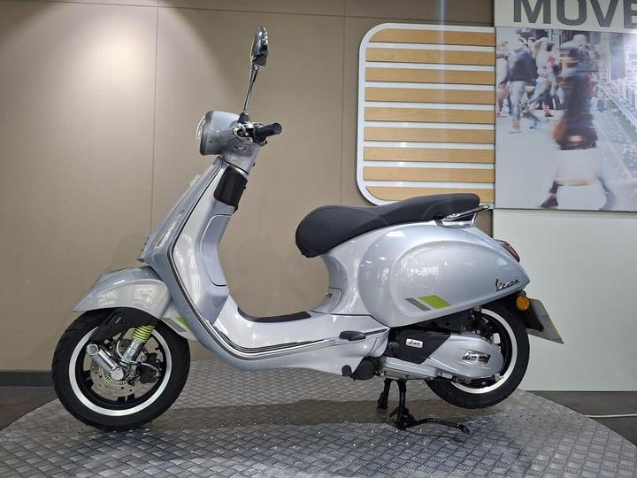 A sleek silver scooter displayed in a showroom, featuring a black seat and modern design elements, set against a neutral background with a blurred image of pedestrians.