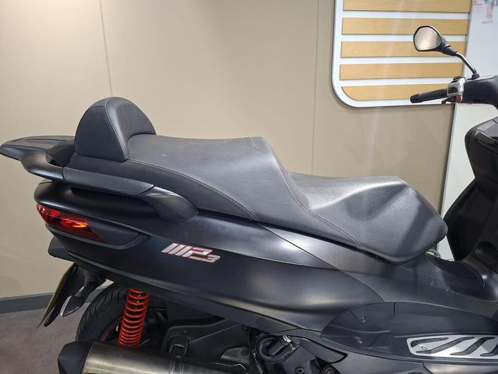 Close-up of a sleek black motorcycle seat with a textured surface, featuring a prominent brand logo and red suspension components in the background.