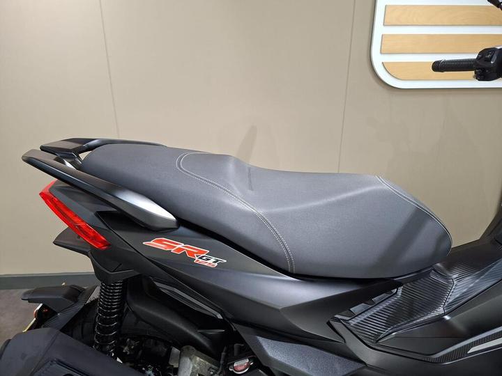 A close-up view of a motorcycle seat featuring a textured black surface with white stitching, positioned on a bike with a sleek design and red accents. The background is neutral, enhancing the focus on the seat and bike details.