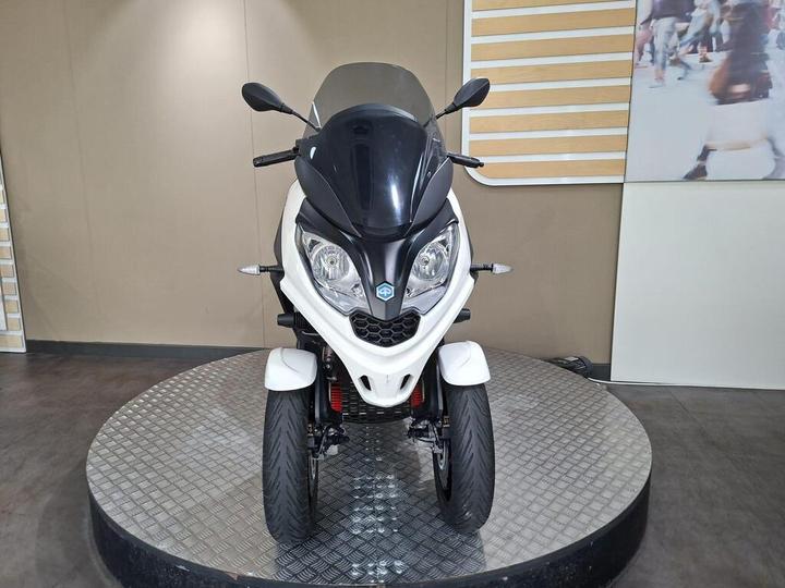 A three-wheeled scooter displayed on a rotating platform, featuring a black and white body, windscreen, and front LED lights. The background includes a beige wall and a panel with images of people.