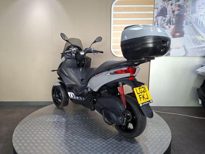 A gray three-wheeled scooter is displayed on a circular platform, with a top case mounted on the rear. The background features a light-colored wall and an abstract image of city life. The scooter has a visible license plate reading "LG21 FKJ."