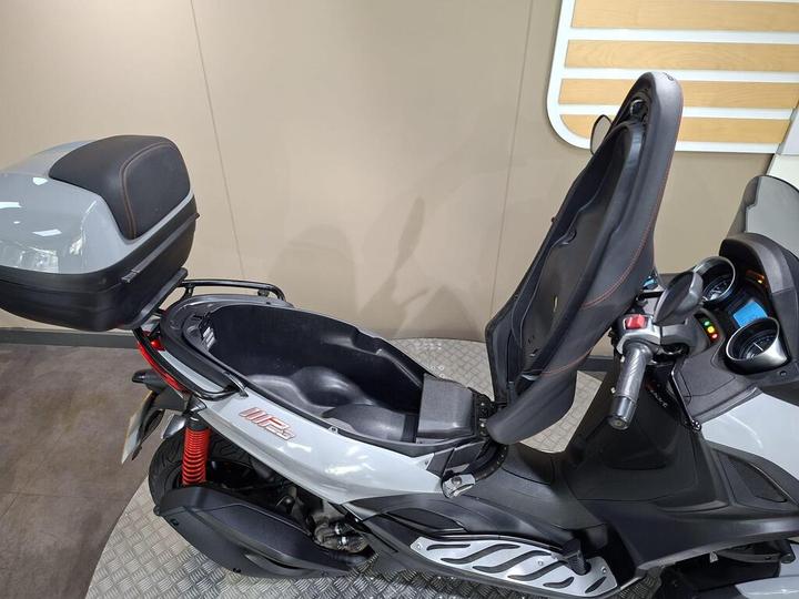 A close-up view of a gray scooter with a high backrest and an open storage compartment. The scooter features a sleek design, a digital display, and a rear trunk mounted on the back. The background is a light-colored wall with wooden accents.