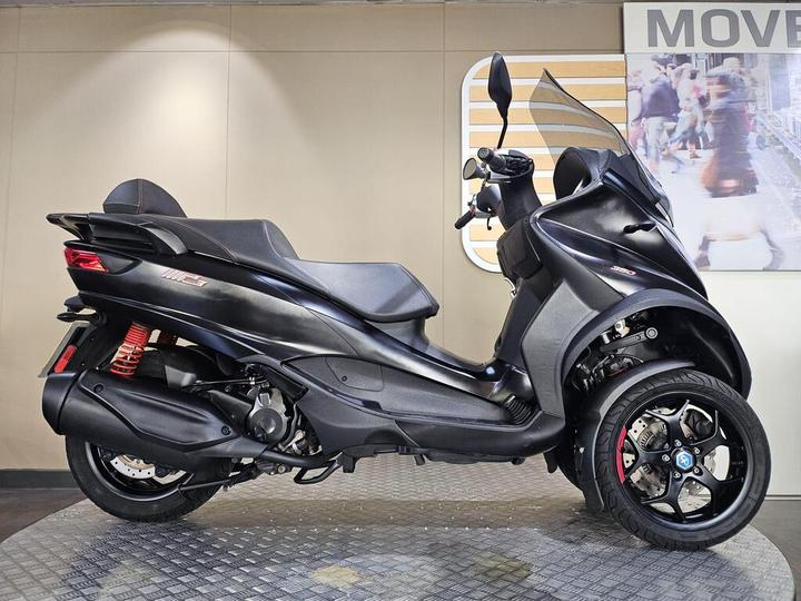 Piaggio MP3 350 (2019) A sleek black three-wheeled scooter is displayed on a textured surface, showcasing its modern design, including a prominent windshield, sporty wheels, and red suspension components. The background features a simple light-colored wall.