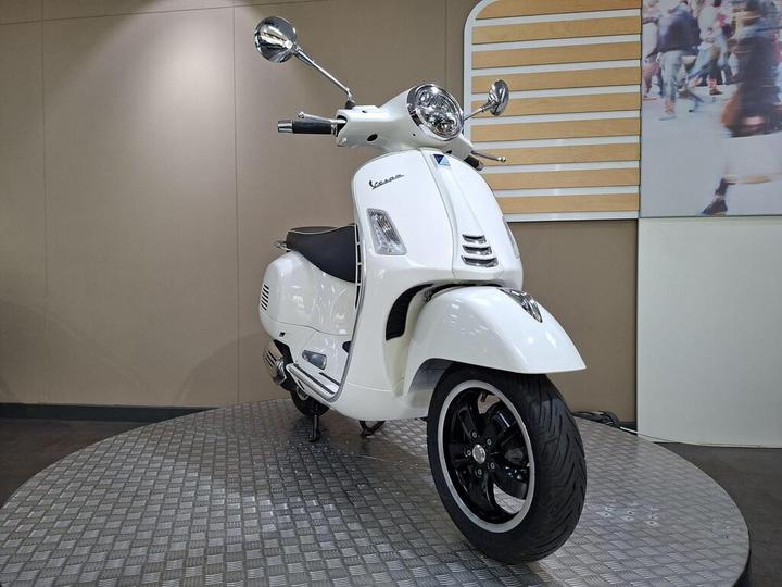A white scooter parked on a textured circular platform with a neutral backdrop.