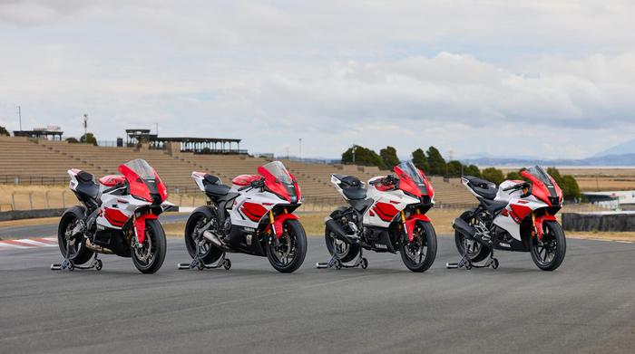 Four Yamaha sport motorcycles are aligned on a racetrack, showcasing a mix of red and white designs. The background features empty grandstands under a cloudy sky.