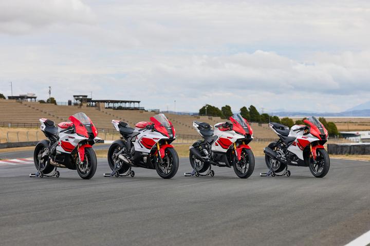 Four Yamaha sport motorcycles are aligned on a racetrack, showcasing a mix of red and white designs. The background features empty grandstands under a cloudy sky.