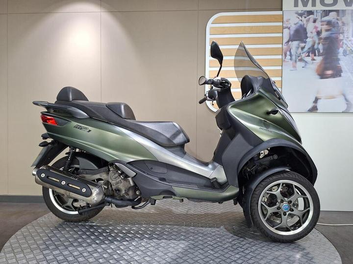 Piaggio MP3 500 LT Sport 2018 A green three-wheeled scooter positioned on a textured platform, featuring a sleek design with a double seat, a windshield, and alloy wheels. The background is a neutral wall with a faint display of pedestrians.